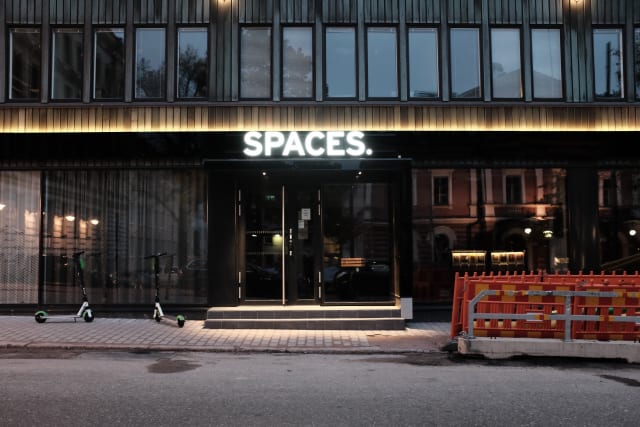 A building in early morning light. Above its door is a lit sign saying 'SPACES.'. Two scooters are on one side of the door, and a construction barrier on the other.