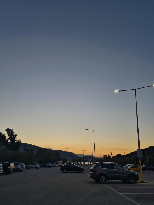 A parking lot with a few cars, as the street lamps come on. The residue orange of a sunset is in the distance.