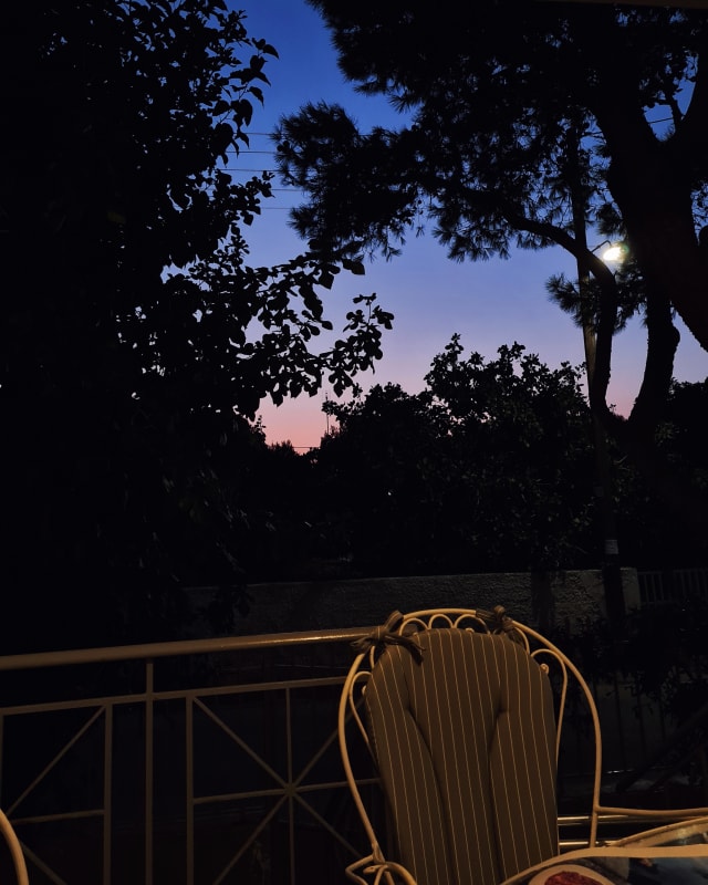 An empty chair on a balcony, with the deep blue or purple of a lingering night behind it.