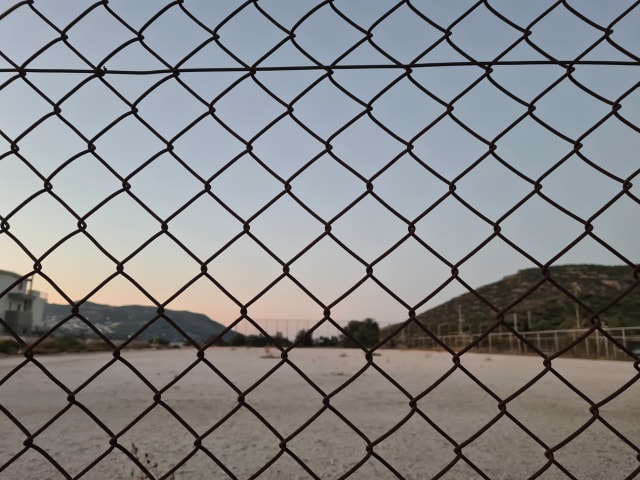 A playing field, empty, behind a fence. The pale blue and pink of a bygone sunset is in the distance.