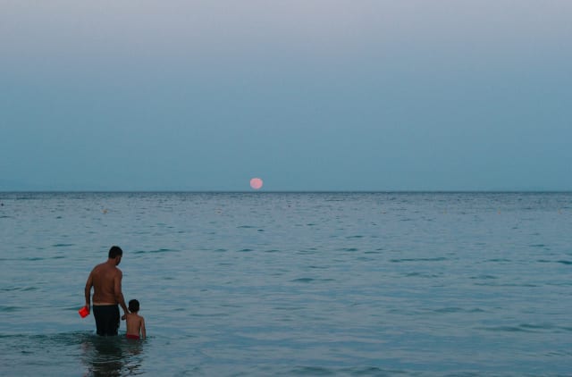 An adult and a child standing in shallow waters, in front of a pale red moon.