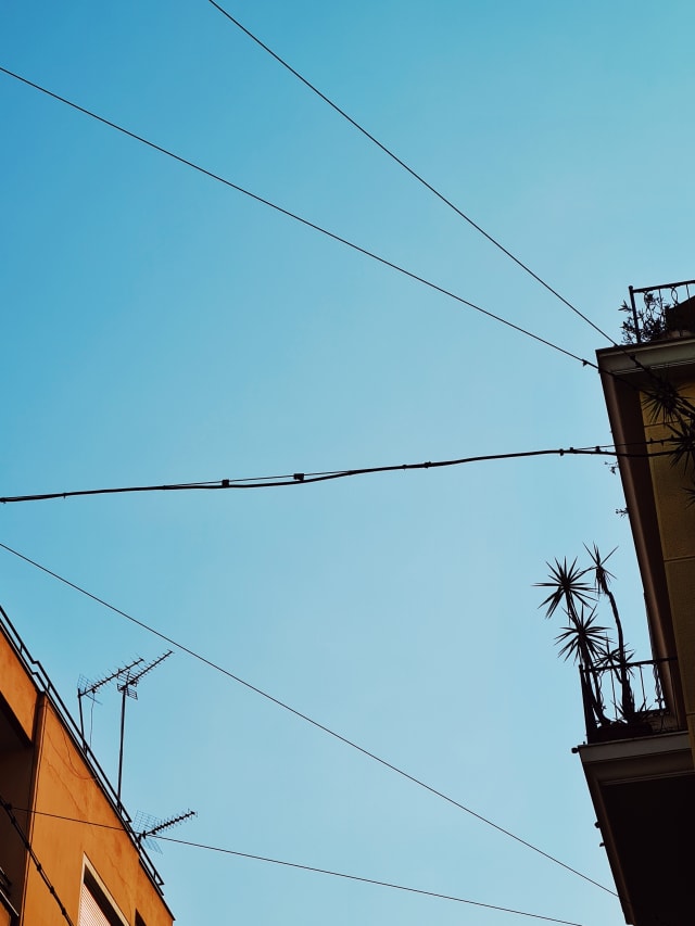 An orange building lit by the sun on one side. On the other side is the darker outline of another building. The blue sky is in the middle and top, with power lines crossing over.