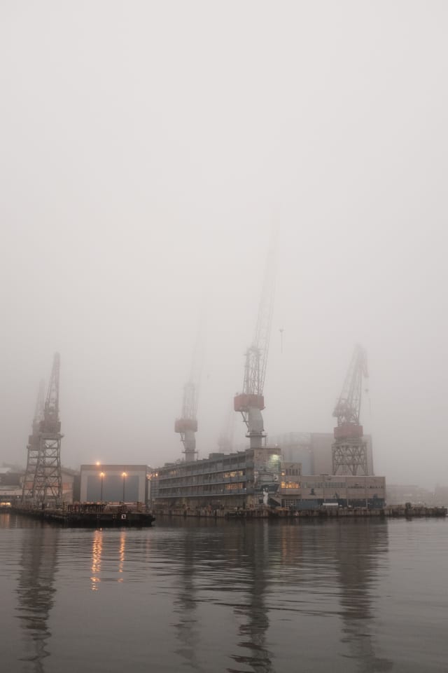 A gray sea in the foreground, with a dockyard in the background. Cranes stand tall and get lost in the fog.