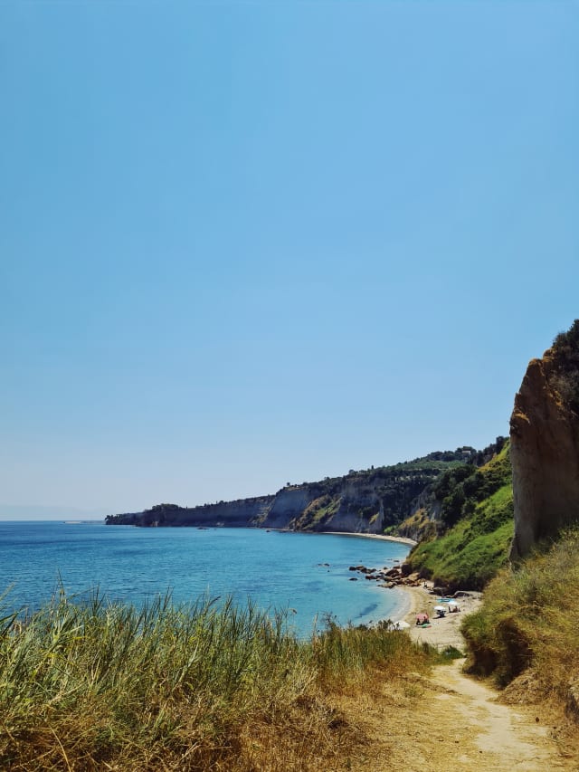 A dirt path with foliage on the side leads to a beach. The sea is bounded by a cove, and has a sparkling blue color.