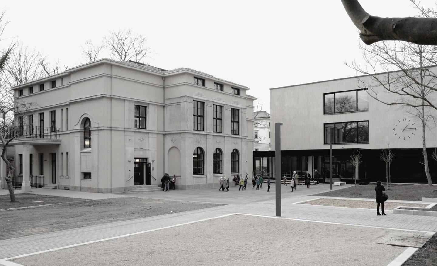 Weißer, historischer Schulbau neben modernem Erweiterungsbau mit großer Fassadenuhr. Kindergruppen auf dem Schulhof zwischen beiden Gebäuden.