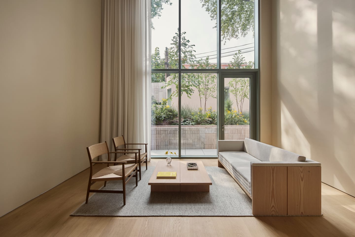 A bright, beige living room with high ceilings and floor-to-ceiling windows with green frames. The natural light draws the eye to the seating area and a large sofa made of light-colored wood.