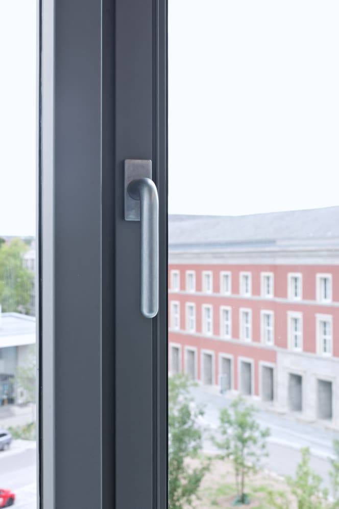 Nahaufnahme des extra angefertigten Fenstergriffs FSB 1147 im Bauhaus Museum Weimar in der Oberfläche Aluminium Pure. Im Hintergrund spielt sich der verschwommene Blick durch das Fenster ab. Ein Haus mit roter Facade.