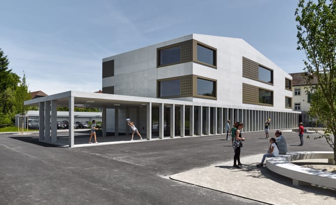 Moderner Schulbau aus hellem Beton mit auskragendem Vordach und Stützenreihe. Kinder und Erwachsene auf dem Pausenhof bei Sonnenschein.