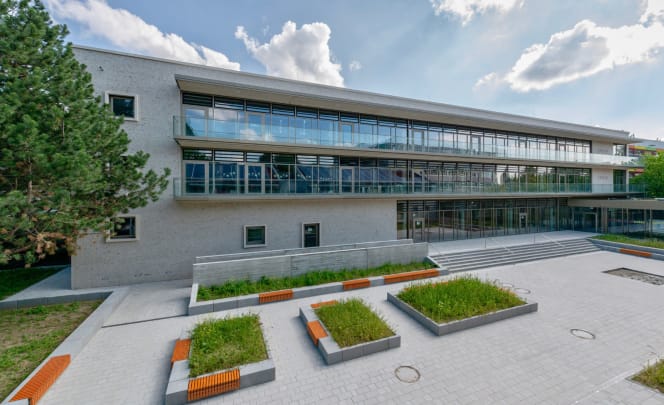 Langer Schulbau mit heller, grauer Fassade und durchgehenden Glasfronten sowie Balkonen aus Glas. Der Pausenhof ist mit Pflanzinseln und Sitzbänken unter einem hellem Wolkenhimmel bestückt.