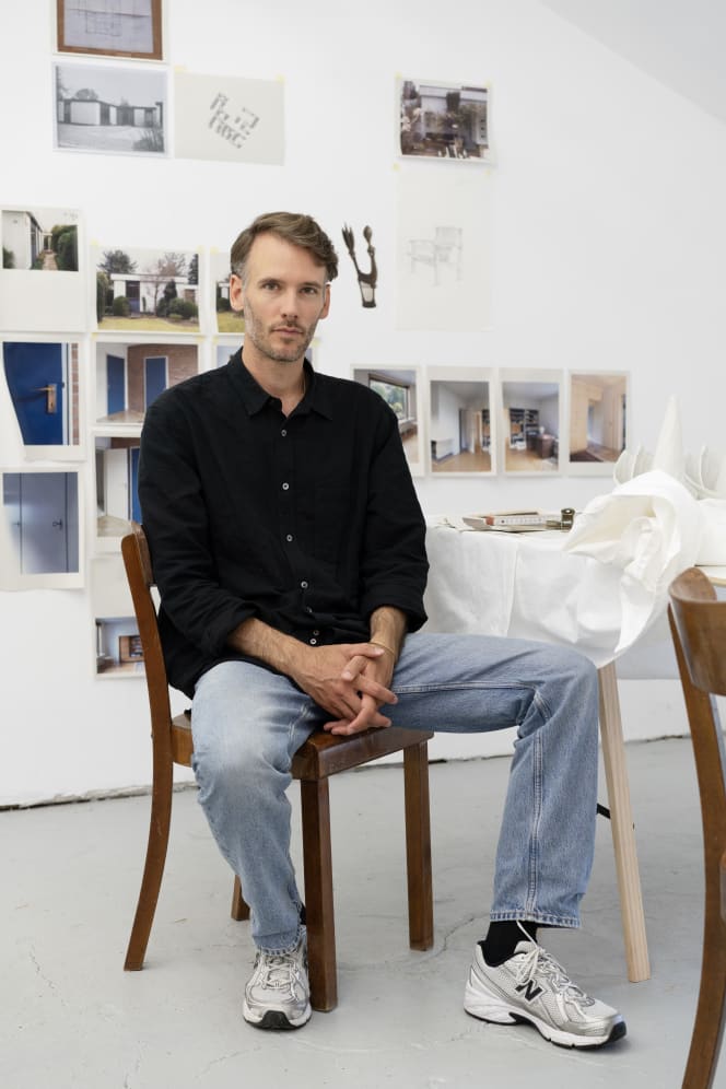 Portrait of Friedemann Heckel in the exhibition space, seated in front of photographs, drawings, and objects.