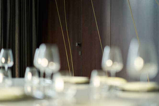 Photo of the FSB 23 0811 door handle in dark bronze-patinated aluminum, seen from a distance. In the foreground, a set dining table with wine glasses appears blurred.