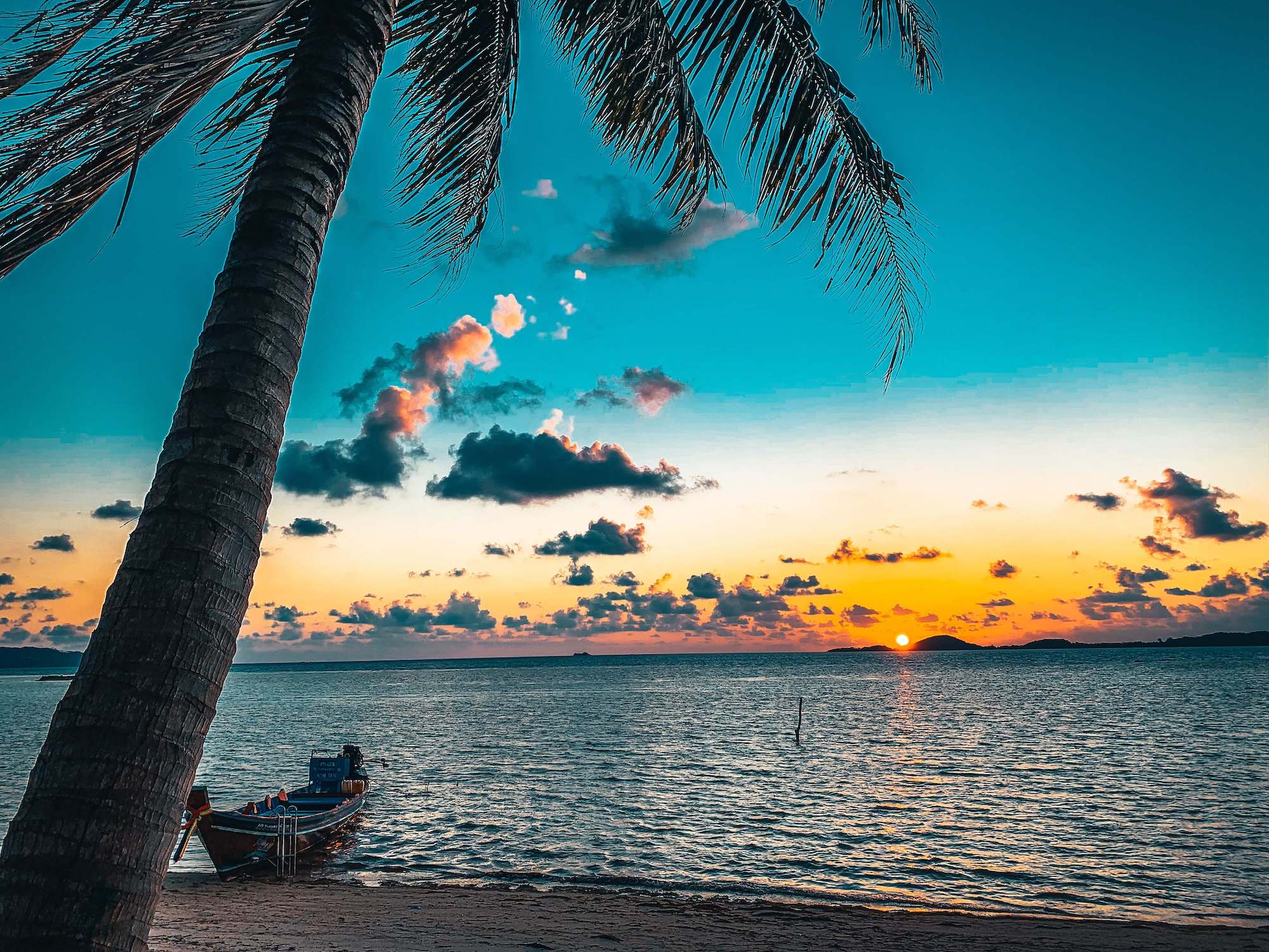 A beach at sunset with the sea and orange sky. There is a boat moored on the beach and a palm tree in view