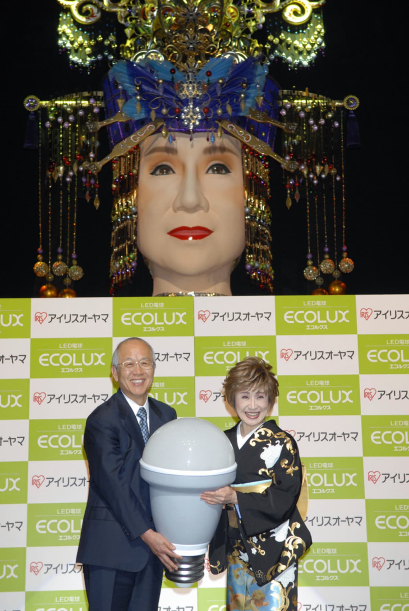 写真は、巨大人形「メガ幸子」の前で、LED電球の新商品を紹介する小林幸子（右）。ニコニコ大百科の説明によると「メガ幸子とは、2009年紅白歌合戦にてその姿を現したラスボス：小林幸子の現時点における最終形態である」（写真：共同通信）