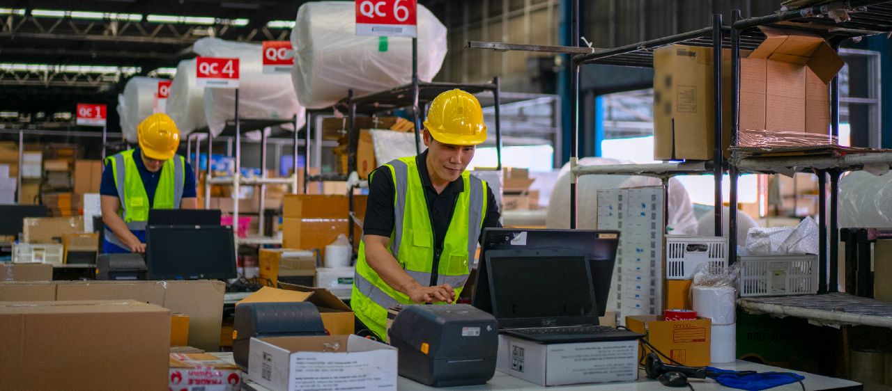 Warehouse workers managing ecommerce orders at a packing station with boxes, laptops, and quality control areas.