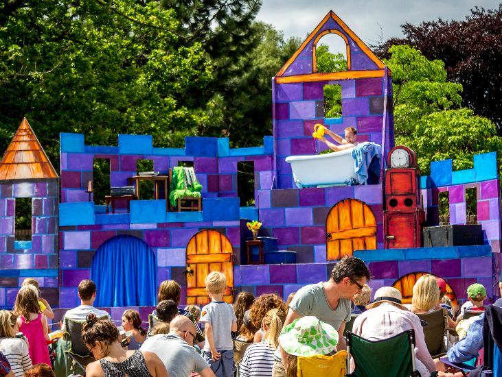 bright blue and purple stage set up for outdoor theatre in the walled garden with actor in a bath tube and people sitting on the grass watching the performance