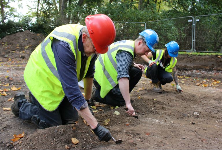 three archaeologists kneeling to dig with their trowels in the dirt