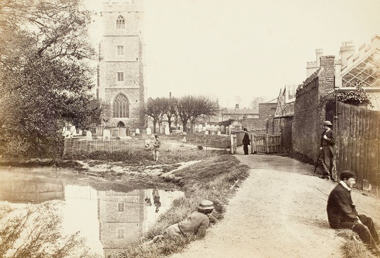 man and child sit beside the moat with all saints church in the distance