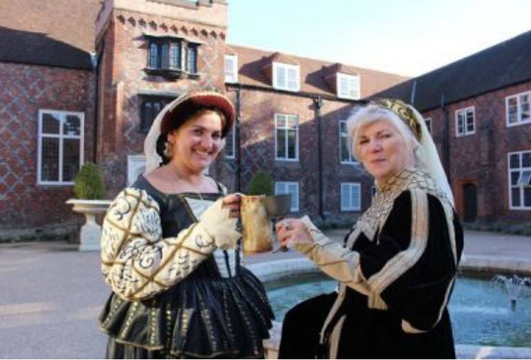 two women dressed in tudor costumes click glasses in the tudor courtyard