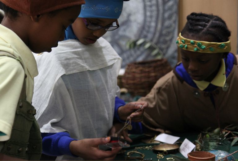Three children dressed in roman costumes look at a roman artefact