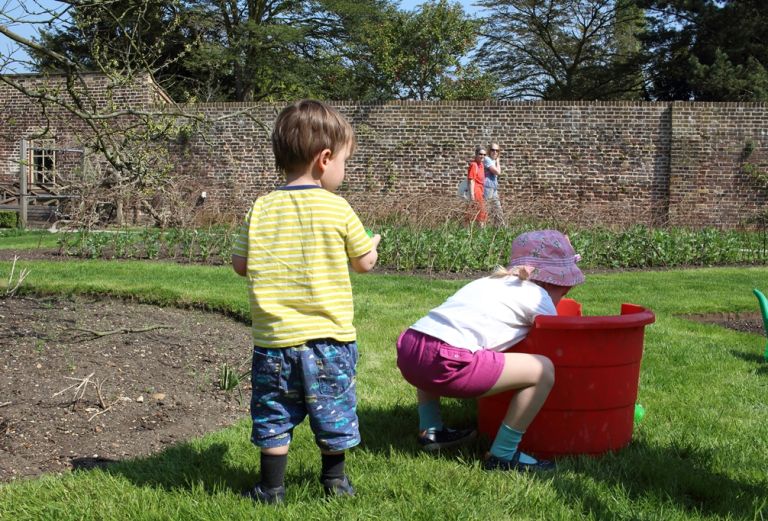 Two children face away from the camera. They are in a garden, and the girl on the right is reaching into a big bucket