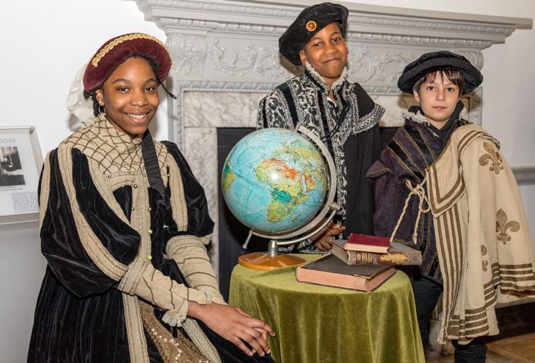 Three children in period dress stand around a table with a globe on it