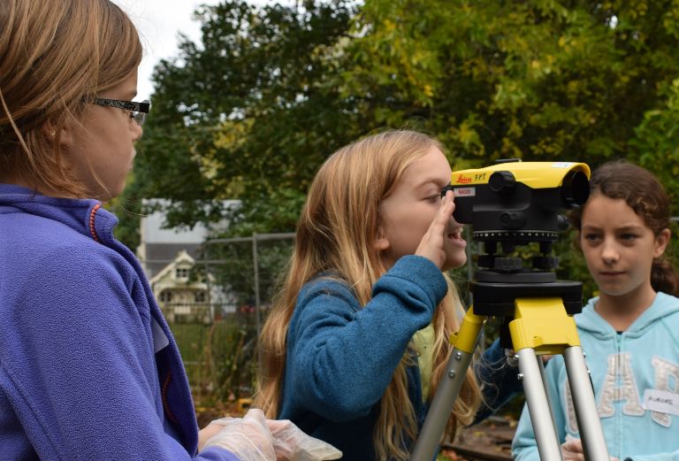 three teenage girls look a surveyor's level on an archaeology dig