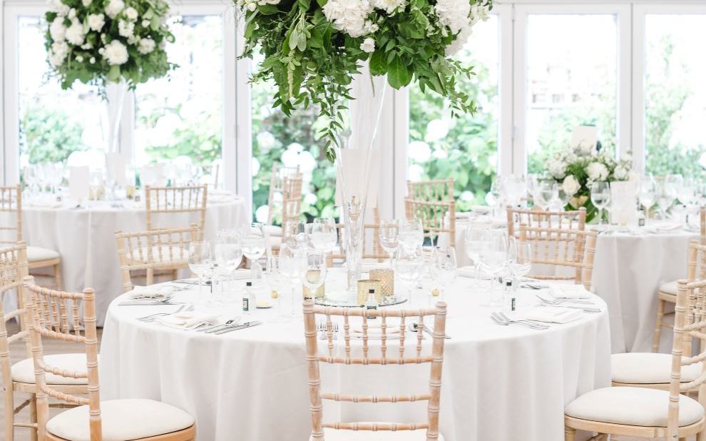Image of table with white cloth, and white chairs. foliage of green leaves and white flowers hangs from the ceiling
