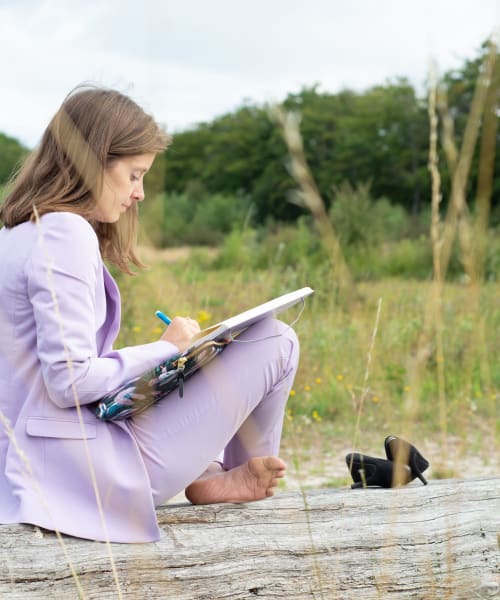Een coachee van het leiderschapstraject voor vrouwen zit in kleermakerszit op een oude boomstronk. Ze schrijft in een schriftje. Ze draagt een lichtpaars pak, haar schoenen liggen naast haar, er is natuur op de achtergrond.
