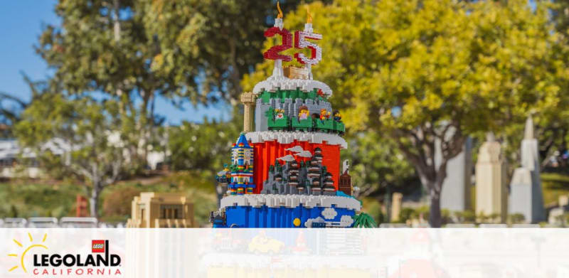 Image features a colorful LEGO cake structure celebrating the 25th anniversary, displayed at LEGOLAND California. The model is set against a blurred background of green trees and a bright blue sky. The LEGOLAND logo is clearly visible at the bottom of the frame.