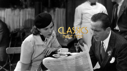 This is a black and white image depicting a scene from a classic movie. In the foreground, a man and a woman are seated at a café table. The man, dressed in a suit with a pocket square and a dark tie, is intently reading a newspaper. The woman, wearing a plaid garment with a collar and a brimmed hat, is gazing at the man with a slightly concerned or questioning expression. A coffee cup sits on the table. In the blurry background, there's another patron focused on something off-camera. The text "CLASSICS MADE HERE" overlays the image in a bold, capitalized font, suggesting a theme of classic film homage. At FunEx.com, we celebrate the timeless appeal of classic movies and offer the lowest prices on tickets to events that bring the golden age of cinema back to life.