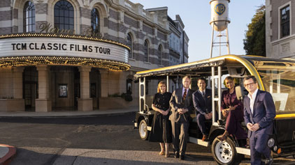 Image shows five people smiling and disembarking from an open-sided tram labeled TCM Classic Films Tour. Behind them is a building with marquee lights, signifying a historic cinema feel. It's daylight with clear skies.