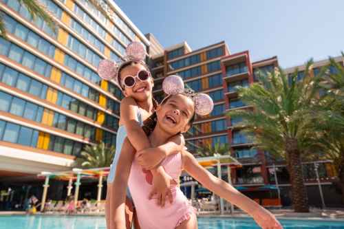 Two children wearing pink outfits and Minnie Mouse ears happily playing by a pool with a colorful hotel building in the background