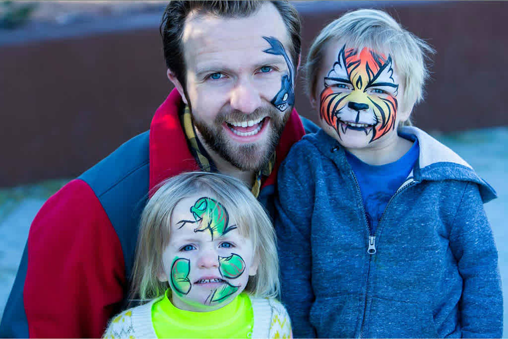 Man and kids with festive face paint smiling outdoors.