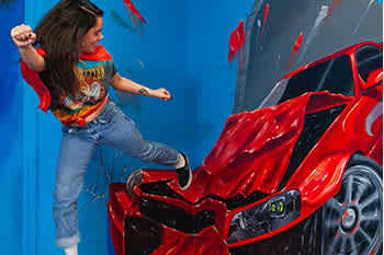 Description: The image features a dynamic scene where a person engages with a three-dimensional artwork that gives the illusion of a shiny red sports car being smashed. The individual appears mid-action, with one leg raised as if stepping out of the car, which is artistically rendered to look as if it's bursting through the wall. The person is wearing a retro graphic tee, denim jeans, and sneakers, with their arm stretched out to balance, enhancing the interactive aspect of the artwork. The background is a simple, blue color, allowing the vibrant red of the car to stand out prominently.

At FunEx.com, we're committed to offering the lowest prices on tickets, ensuring you can enjoy these incredible experiences without breaking the bank.