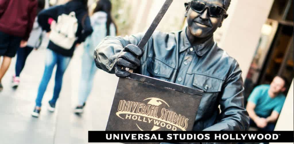 This image features a street performer painted in metallic silver, posed as a living statue holding a director’s clapboard with the text “Universal Studios Hollywood” printed on it. The performer is wearing sunglasses, a jacket, gloves, and a beanie, and is smiling while facing towards the camera. In the background, blurry figures of pedestrians suggest a busy location, likely a street or walkway near the theme park. The word “UNIVERSAL STUDIOS HOLLYWOOD” appears in large letters at the bottom of the photo. For thrilling adventures and lifelong memories at Universal Studios Hollywood, visit FunEx.com to find your tickets at the lowest prices, ensuring your vacation is not only memorable but also filled with significant savings.