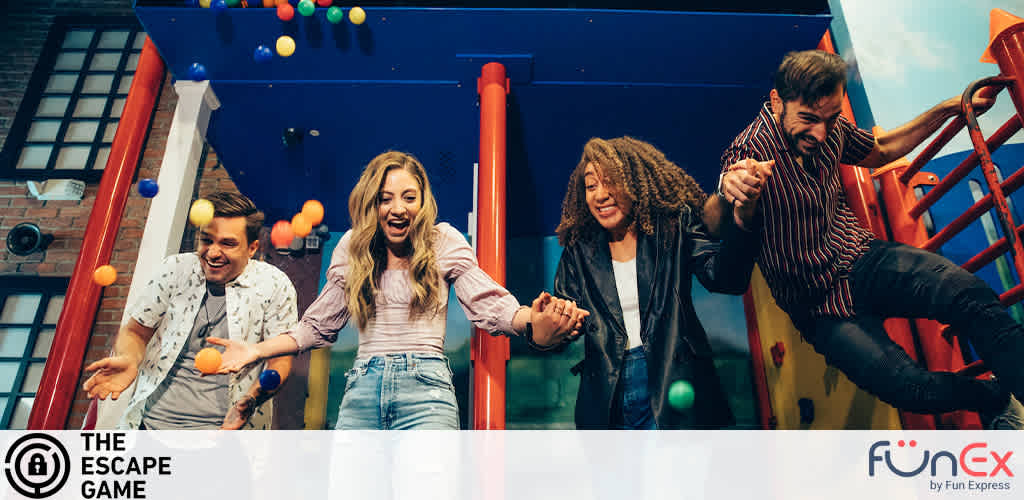 Four joyful friends are exiting a colorful playground slide amidst a shower of multi-colored balls. They're all smiling and appear to be enjoying themselves. The playful setting has a backdrop suggestive of an urban landscape. Logos for The Escape Game and FunEx are visible, indicating a fun, gaming experience.