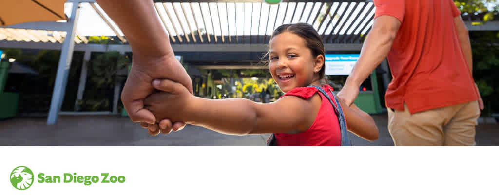 A joyful child in a red top holds hands with an adult, gazing back with a bright smile, at the entrance of the San Diego Zoo. Sunshine and greenery set a cheerful mood. The zoo's logo is visible in the corner.