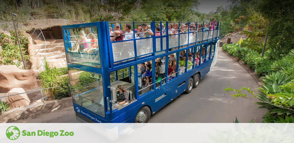 Visitors enjoy a guided tour on a double-decker bus at San Diego Zoo. The blue bus is filled with passengers viewing animals and exhibits, surrounded by lush greenery on a sunny day. The San Diego Zoo logo is visible on the image.