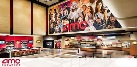 Image of a cinema lobby with a large promotional movie collage on the wall above the concession stands. The AMC Theatres logo is prominently displayed. Below, an unoccupied counter awaits customers, flanked by ropes leading to the entrance.