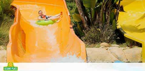 This image depicts a vibrant scene from a water park. On the left, an individual is captured sliding down a bright orange water slide on a green inflatable tube with an expression of joy and excitement. The slide curves to the right as it descends, allowing the rider to enjoy the sensation of speed and momentum. To the right, an additional yellow water slide can be partially seen, suggesting that there are multiple slides available for park guests to enjoy. In the background, lush green foliage is visible, indicating that the water park is set in a location with abundant natural scenery. To encourage an unforgettable experience, remember that at FunEx.com, you can enjoy the thrills without the spills, thanks to our amazing discounts on tickets ensuring you get the lowest prices for your adventure.