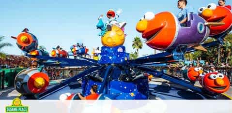 Colorful amusement park ride featuring Sesame Street characters. Each ride car, designed like different characters such as Elmo and Cookie Monster, spins around a central axis. Children and adults sit inside, enjoying the motion under a sunny sky with lush greenery in the background. The Sesame Place logo is visible in the corner.