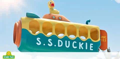 Image of a large, whimsical boat named S.S. Duckie. It's designed to look like a floating bathtub toy with vibrant yellow, green, and orange colors. A joyful rubber duck sits on top, and the hull has round, open portholes. The sky in the background is a clear blue. There's a small emblem reading 'Sesame Place' at the lower left corner.