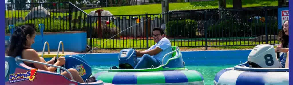 Visitors enjoy an outdoor bumper boat attraction on a sunny day. People are seated in colorful individual boats with large inflatable rings. They're navigating a small blue pool, surrounded by a green fence and grassy areas. Some guests are steering while talking or laughing with each other.