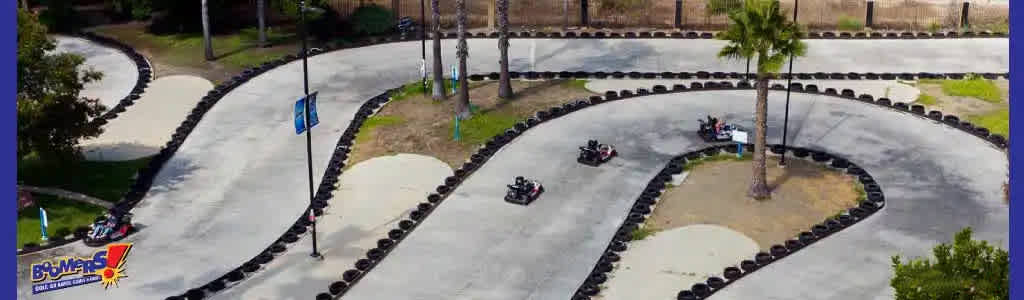 Aerial view of an outdoor go-kart track with winding asphalt lanes bordered by tires. Three go-karts navigate the course on a sunny day amid greenery and a few palm trees. A blue handicap parking sign is visible near the track.