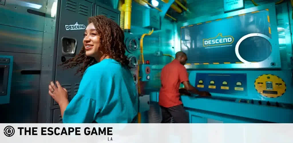 This image features a brightly lit, room designed to resemble the interior of a submarine with various shades of blue and yellow trim. A woman in a turquoise sweater is seen smiling at the camera, her curly hair framing her face, as she looks over her shoulder while gripping a handle on a dark metallic door with the word "DESCEND" written above it. In the background, a man in a red shirt is focused on interacting with a console that has an array of buttons and a large, circular window to the side. Overhead, signage that reads "DESCEND" matches the theme of the room, and a logo indicating "THE ESCAPE GAME LA" is visible in the bottom left corner of the image. Enjoy your next adventure with FunEx.com and unlock savings with our lowest prices on tickets for memorable experiences.