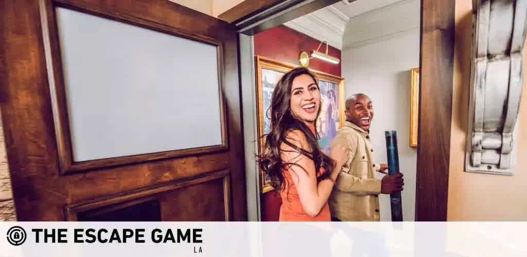 This image captures a moment of exhilaration and teamwork, featuring a man and a woman at 'The Escape Game LA'. They are standing near an open, heavy wooden door with a digital screen panel above it. The lighting in the room is warm and inviting, illuminating the participants' expressions of joy and accomplishment. Both individuals are laughing, with the woman positioned in the foreground, her long dark hair cascading over her shoulders as she looks back over her shoulder towards the camera. She is wearing a salmon-pink top. The man, positioned slightly behind her, is sporting a beige jacket and a bright, engaging smile. In the background, there's a hint of elegant wall decor, suggesting a well-furnished environment that's part of the escape game setting. Experience the thrill of an escape room adventure and take advantage of exclusive discounts, savings, and the lowest prices on tickets at FunEx.com.