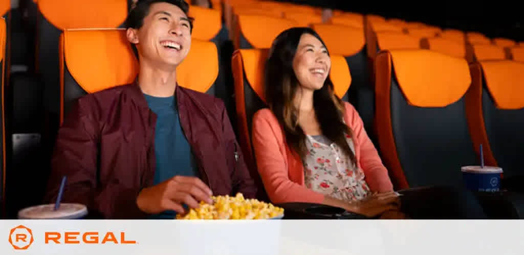 Image shows a cheerful man and woman sitting in a movie theater with orange seats. The man, on the left, wears a burgundy jacket over a teal shirt, holding a popcorn bucket. The woman, on the right, smiles widely, wearing a pink cardigan over a flower-patterned top. A soda cup rests in her cup holder. The Regal Cinemas logo is visible at the bottom. They appear to be enjoying a film.