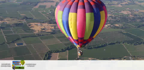 This image shows a colorful hot air balloon in mid-flight, viewed from a slight elevation. The balloon features a pattern of vertical stripes in vibrant colors such as red, yellow, blue, green, and purple, and is topped by a dark blue cap. It is floating over a vast landscape dotted with patches of agricultural fields, which form a patchwork of different muted green and brown hues. The sky is clear, indicating favorable weather conditions for ballooning. The horizon stretches across the background, emphasizing the expansive view from the balloon. Experience the thrill of a hot air balloon ride and make memories to last a lifetime – be sure to check FunEx.com for the lowest prices and tickets, ensuring you get the best savings on your next adventure.