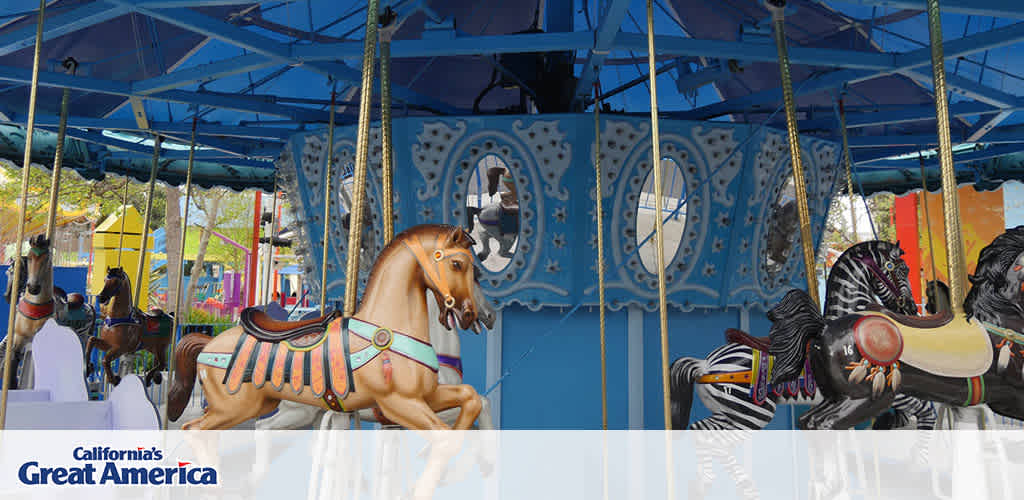 Image features a colorful carousel at California's Great America. A variety of traditional carousel horses and a zebra figure are shown with intricate designs, poised mid-gallop. Some are brown, others are white with colorful markings, all adorned with bright saddlery. The ride's structure includes a blue canopy above and decorative panels around the center, reflecting classic fairground artistry. A glimpse of the park's environment is visible in the background.