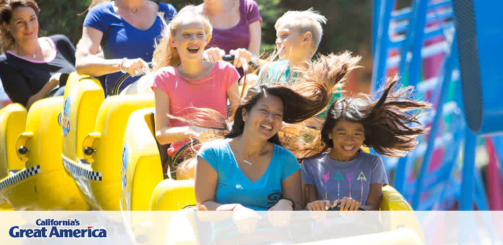 This image features a diverse group of individuals experiencing a thrilling roller coaster ride at California's Great America. There are multiple rows of bright yellow coaster seats filled with passengers. The focus is on two rows, with a woman and a young girl in the front row, both smiling broadly and enjoying the ride. The young girl has her hair dramatically swept back by the wind, indicating the high speed of the roller coaster. There are trees and a clear blue sky in the background, as well as a part of a coaster's blue structural element. The sunshine and the expressions of joy on the riders' faces convey a sense of excitement and fun typical of an amusement park setting.

Experience the exhilaration of California's Great America and enjoy the savings when you purchase your tickets through FunEx.com, where we strive to offer the lowest prices available.